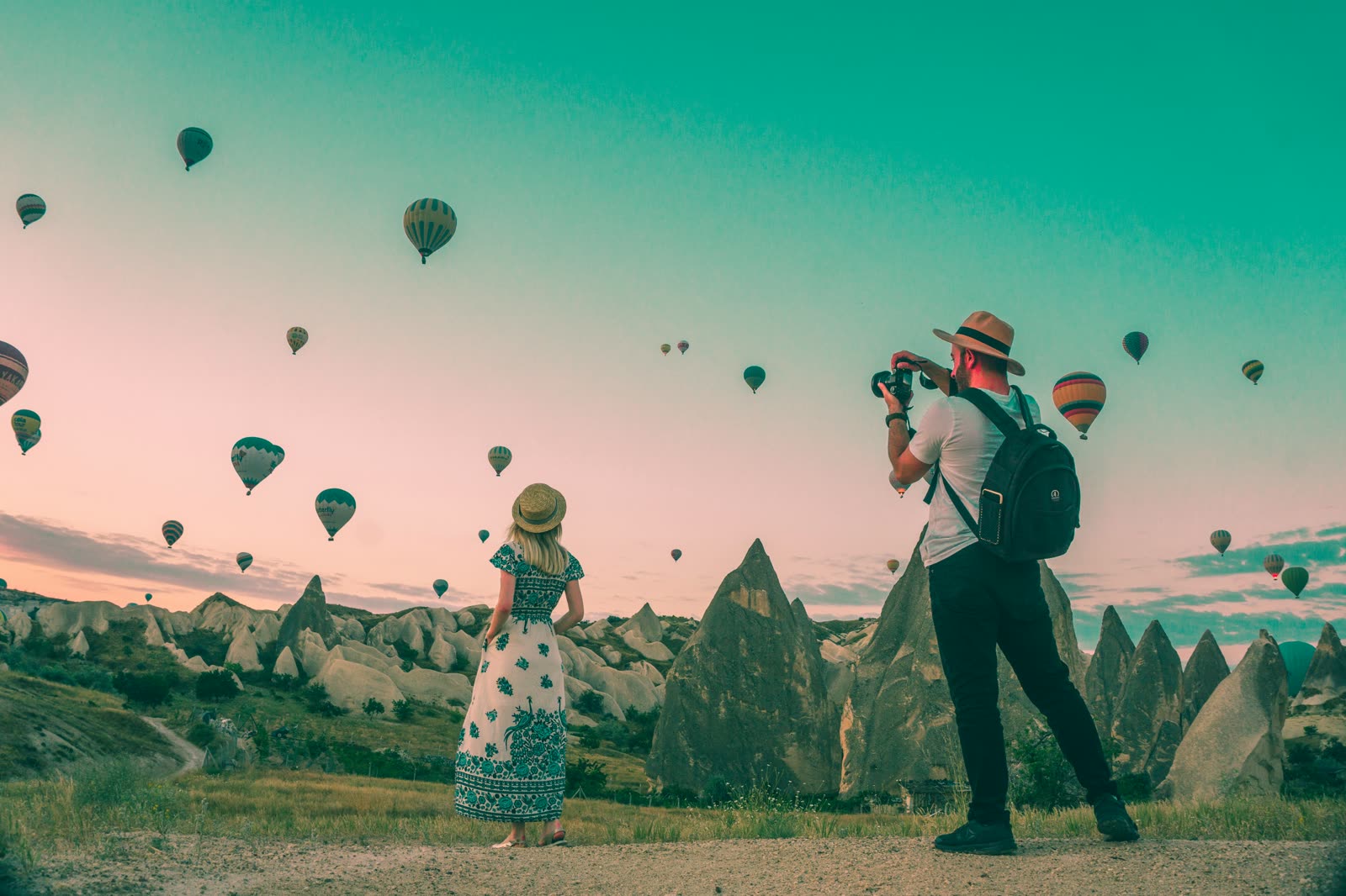 Two travelers watching hot-air balloons rise over a valley at dawn